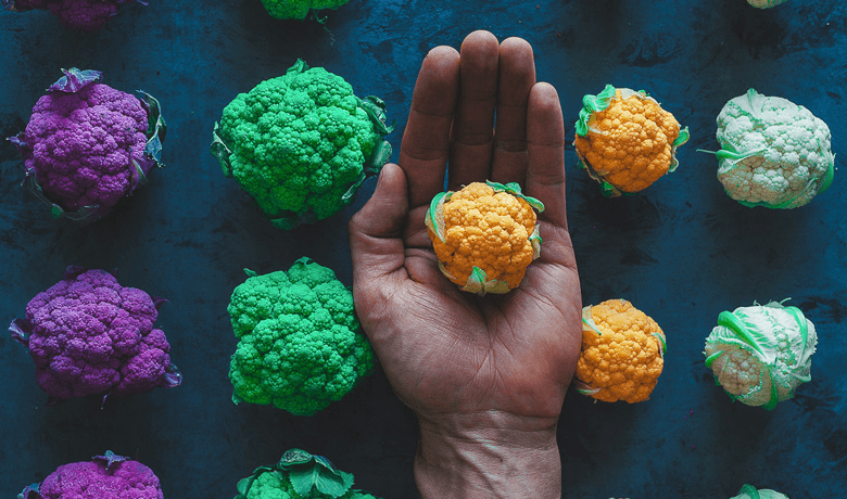 hand holding out orange colored cauliflower against a dark background with rainbow cauliflower lined up from left to right in columns purple, bright green, orange and pale green