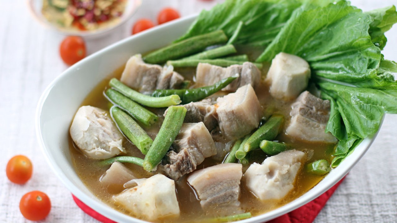 white bowl with sour filipino broth, green leaves and cubes of pork belly